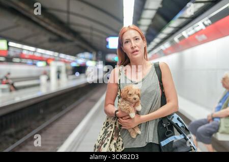 Une femme tient un petit chien dans une gare. Le chien est brun et c'est un caniche. La femme porte un sac et un sac à dos, et il y en a d'autres Banque D'Images