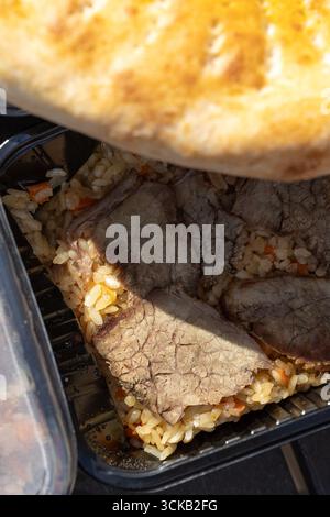 Gros plan de riz pilaf d'agneau avec des tranches de viande tendres, des carottes et du pain plat doré - repas copieux et savoureux dans un récipient en plastique, parfait pour le takeou Banque D'Images