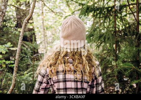 Vue arrière d'une fille aux cheveux blonds bouclés, portant un chapeau et une veste à carreaux, debout dans la forêt. Temps ensoleillé. Banque D'Images