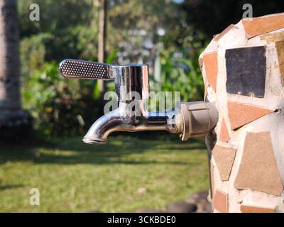 Robinet d'eau extérieur brillant fixé à un mur décoré de carreaux de céramique. Le fond montre un jardin avec de l'herbe verte et des plantes Banque D'Images