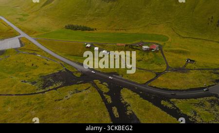 Image aérienne d'une zone rurale islandaise avec une maison au toit rouge, des champs verts, un sol volcanique, une route sinueuse, des véhicules, et des collines ondulantes. Banque D'Images
