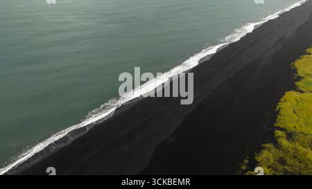 Perspective aérienne d'une plage de sable noir rencontrant l'océan, avec des vagues de mousse blanche et une végétation verte vibrante contrastant avec le terrain volcanique. Banque D'Images