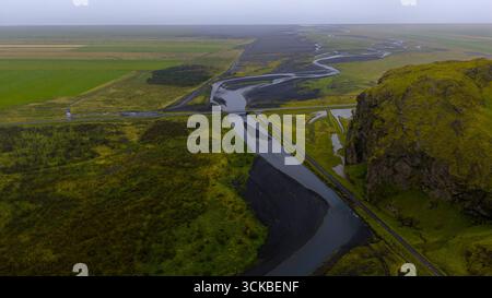 Une rivière sinueuse coule à travers un sol volcanique noir et des champs verts en Islande. Un petit pont et une colline rocheuse couverte de mousse sont visibles sous un overc Banque D'Images