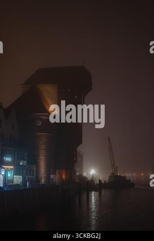 La grue Zuraw illuminée à Gdansk, en Pologne, se dresse contre un ciel nocturne brumeux. Les reflets scintillent sur la rivière, avec une grue lointaine en vue. Banque D'Images