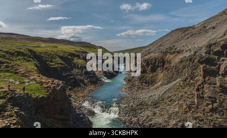 Une rivière bleue éclatante coule à travers un canyon accidenté en Islande, avec une végétation verte, des falaises rocheuses, une petite cascade, et des gens mettant en valeur le sca Banque D'Images