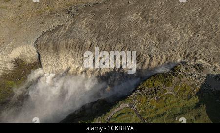 Image aérienne d'une cascade en Islande, avec de l'eau boueuse en cascade dans la brume, entourée de mousse verte et de terrain rocheux. Un arc-en-ciel apparaît dans le mis Banque D'Images