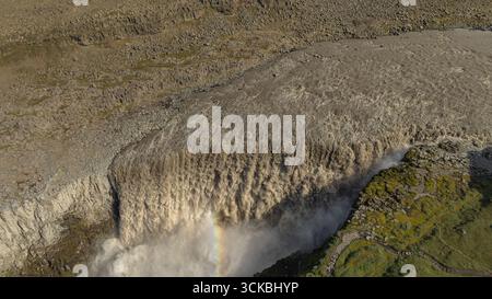 Vue aérienne spectaculaire de la cascade Dettifoss avec de la brume, un arc-en-ciel faible, un terrain rocheux, une végétation verte et des sentiers de randonnée visibles en Islande. Banque D'Images