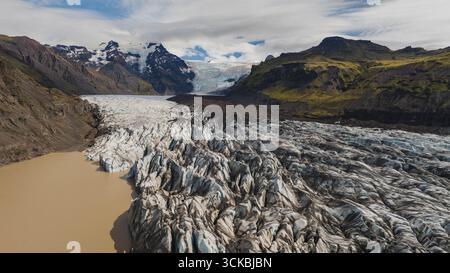 Vue aérienne spectaculaire d'un glacier en Islande qui coule vers un lac glaciaire laiteux, encadré par des montagnes enneigées escarpées et des pentes verdoyantes. Banque D'Images