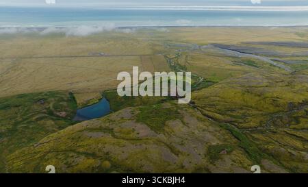 Image aérienne de collines couvertes de mousse, d'un étang bleu, de ruisseaux sinueux, de plaines dorées et de l'océan rencontrant l'horizon sous des nuages bas en Islande. Banque D'Images