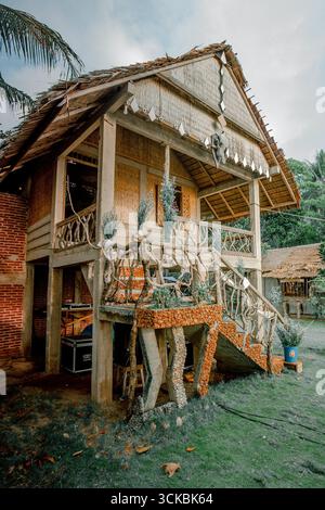 Une maison rustique en bois avec un toit de chaume, avec des rambardes d'arbres uniques et un escalier, entouré de verdure Banque D'Images