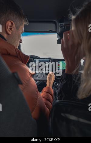 Deux individus sont assis dans un véhicule, l'un utilisant un contrôleur de drone avec une vue aérienne en direct sur l'écran. Le tableau de bord et le paysage couvert sont visibles. Banque D'Images
