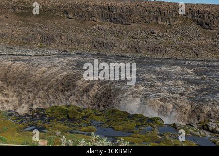 Les cascades de Dettifoss avec une immense eau boueuse sur des falaises accidentées, entourées de terrains rocheux, de mousse verte et de formations de basalte. Banque D'Images
