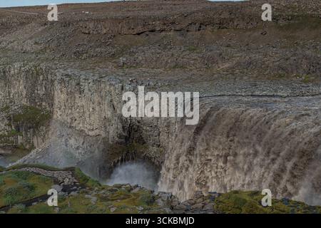 La cascade de Dettifoss cascades sur des falaises de basalte sombres, entourées d'un terrain rocheux avec des taches vertes. La brume monte comme un chemin et la zone de visualisation sont visibles Banque D'Images