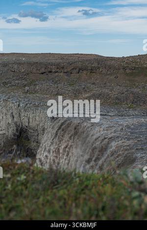 La cascade de Dettifoss dans le parc national de Vatnajokull cascades au-dessus de roches volcaniques sombres, avec de la brume qui monte et un ciel partiellement nuageux au-dessus du terrain aride. Banque D'Images