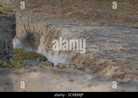 Dettifoss cascade avec une force immense dans une gorge brumeuse, entourée de falaises rocheuses et de mousse, avec de minuscules personnages visibles sur le bord de la falaise. Banque D'Images