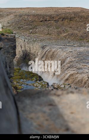 Dettifoss cascades au-dessus d'une falaise accidentée avec la brume qui monte. Le terrain rocheux, la végétation clairsemée et les personnages lointains soulignent l'immense échelle de la cascade Banque D'Images