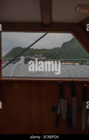 Scène de l'intérieur d'un bateau montrant un panneau de bois avec des couteaux, un fjord calme, des maisons colorées sur un rivage, des collines verdoyantes et un ciel couvert. Banque D'Images