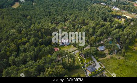 Une zone boisée luxuriante à Nowy Lubiel, en Pologne, avec une maison blanche avec un toit rouge, des structures plus petites, une route et une dense verdure environnante. Banque D'Images