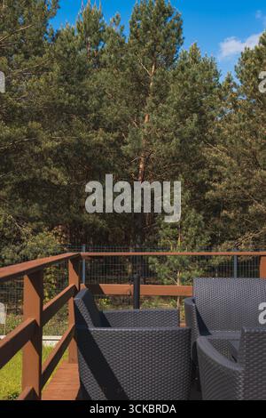 Une terrasse en bois présente des chaises en osier foncé autour d'une table, bordée par une balustrade et une clôture en treillis métallique, avec une forêt de pins dense en arrière-plan. Banque D'Images