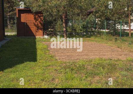 Une cour arrière ensoleillée avec une parcelle de sol labouré, un hangar en bois et une clôture en fil vert. Les pins et les bâtiments voisins sont visibles en arrière-plan. Banque D'Images
