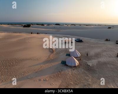 Vue aérienne des tentes nichées parmi les dunes de Steroh, où le sable doré rencontre la mer Azur sous un soleil doux et délavé, dunes de Steroh, Socotra, Yémen. Banque D'Images