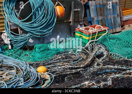 L'image montre un enchevêtrement coloré de filets de pêche, de cordes et de bouées empilés sur le sol à côté de caisses en bois et d'équipements métalliques altérés. Th Banque D'Images