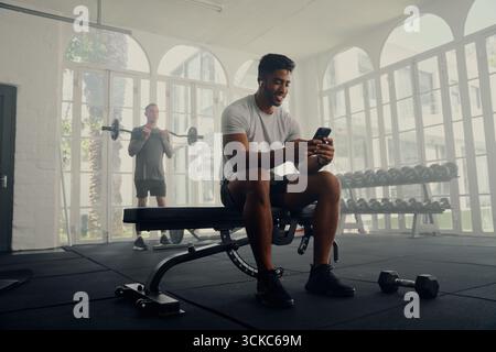 Deux jeunes hommes multiraciaux vêtus de vêtements de sport, exerçant et utilisant un téléphone portable au gymnase Banque D'Images
