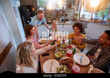 Grand groupe de jeunes adultes multi éthiques déjeunant ensemble dans une cuisine domestique, grillant avec des boissons près de la table. Maison, convivialité, divertissement Banque D'Images