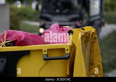 Gros plan d'une poubelle jaune remplie d'un sac poubelle rouge, avec un camion de collecte des déchets en arrière-plan. L'image illustre la gestion des déchets urbains Banque D'Images
