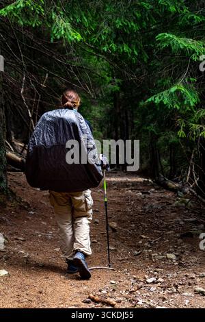 Fille avec un grand sac à dos de randonnée recouvert d'une couverture de pluie et de bâtons de trekking marchant sur le sentier à travers la forêt de conifères sombre. Photo verticale Banque D'Images