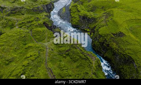 Une rivière sinueuse coule à travers une vallée verdoyante en Islande, entourée de falaises abruptes, d'affleurements rocheux et de sentiers visibles à flanc de colline. Banque D'Images