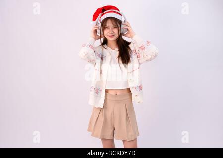 souriante belle femme célébrant noël portant chapeau de père noël et écouteurs en fond blanc de studio Banque D'Images