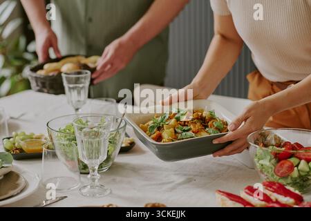 Jeune femme caucasienne servant la salade tandis que jeune homme préparant la nourriture à table Banque D'Images
