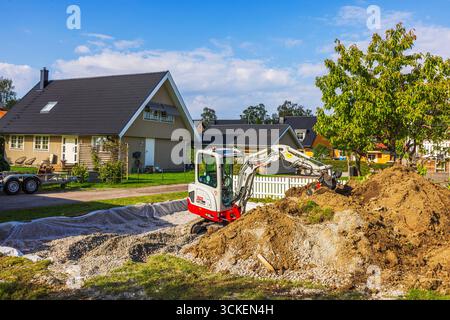 Mini pelle excavatrice creusant du sol près des villas par une journée ensoleillée avec du gravier et des matériaux de construction sur le sol. Suède. Stockholm. Banque D'Images