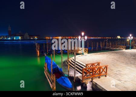 Romantique vue nocturne de Venise avec des gondoles amarrées le long du front de mer éclairé. Les lampadaires lumineux et les eaux calmes créent une atmosphère sereine Banque D'Images