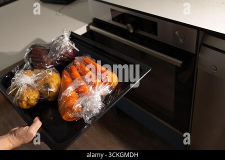 Préparer un repas sain, une main insère un plateau de légumes enveloppé dans des manchons de cuisson dans le four pour une méthode de cuisson alternative dans une cuisine domestique. Banque D'Images