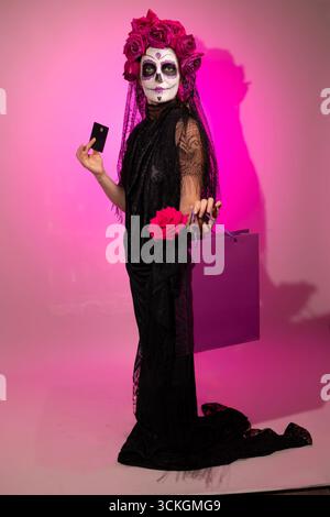 Une fille portant un costume de calavera catalina avec de la peinture de visage de crâne de sucre, une couronne de fleur, et une robe noire se tient debout avec une rose, un sac à provisions, et une carte de crédit sur un fond rose. Banque D'Images