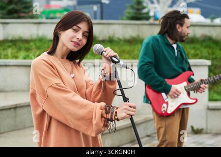 Portrait d'une jeune femme caucasienne chantant dans un microphone à l'extérieur tandis que jeune homme caucasien jouant de la guitare électrique en arrière-plan, les deux jouant de la musique dans un environnement de parc urbain Banque D'Images
