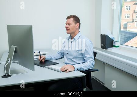 Homme d'affaires danois au travail à côté de son ordinateur assis dans la chemise bleue dans le bureau souriant, homme de travail dans la chemise bleue dans le bureau blanc Banque D'Images