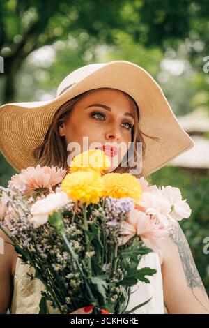 Femme élégante tenant un bouquet de fleurs portant un chapeau de soleil dans le parc Banque D'Images