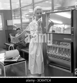 Distributeur automatique 1957. Une femme blonde est appuyée contre le distributeur automatique qui offre des barres de chocolat de différentes sortes. La jupe complète typique populaire pour les femmes en 1957 était une large jupe évasée En A ou en cercle, souvent serrée à la taille avec une ceinture pour créer une silhouette de sablier. Fabriqué à partir de tissus comme le coton, le tulle ou la mousseline, il est généralement tombé à mi-mollet et a été porté avec des jupons pour plus de volume. Le style était vibrant, féminin et emblématique de la mode 'New look' des années 1950. Conard ref 3496 Banque D'Images