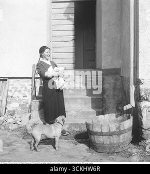 Une femme âgée se tient près des escaliers avec de l'épicerie et des bouteilles de lait dans ses bras. Un chien se tient à ses côtés. Une baignoire en bois usée est placée sous le tuyau d'eau de pluie. 1954 Kristoffersson réf. BX32-5 Banque D'Images