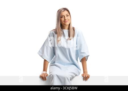 Jeune patiente dans une robe d'hôpital assise sur un panneau blanc isolé sur fond blanc Banque D'Images