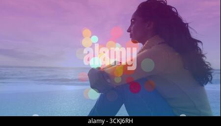 Femme assise portant un pull serrant les genoux sur la plage au crépuscule, cercles bokeh translucides, espace de copie Banque D'Images