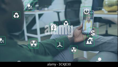 Tenant smartphone homme afro-américain visionnant l'appel virtuel sur le sol du bureau, avec des icônes d'énergie Banque D'Images