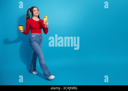 Charmante jeune femme tenant un smartphone avec une tasse à café, souriant avec confiance sur un fond bleu vif Banque D'Images