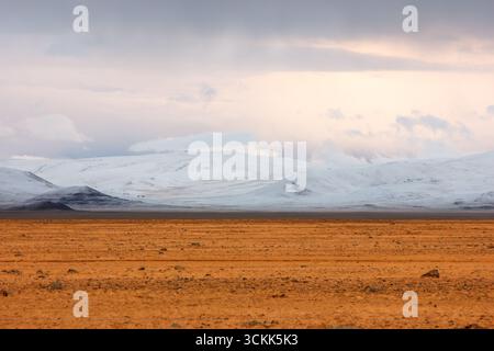 Vue panoramique d'une vaste plaine désertique d'automne avec des sommets enneigés contrastés et un ciel nuageux spectaculaire, créant une atmosphère surréaliste et contrastée Banque D'Images