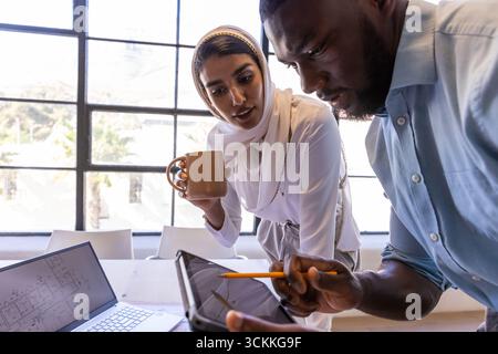 Divers collègues se penchant sur le bureau examinant les plans avec ordinateur portable, tablette, tasse à café Banque D'Images