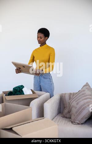 Femme afro-américaine debout dans le salon par canapé déballant des boîtes et examinant des œuvres d'art encadrées Banque D'Images