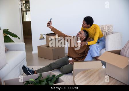 Couple diversifié prenant selfie avec smartphone dans le salon parmi les boîtes tout en tenant des tasses à café Banque D'Images